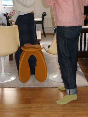Person wearing dark jeans and green socks standing in a living room in front of a yellow stool on a wooden floor.